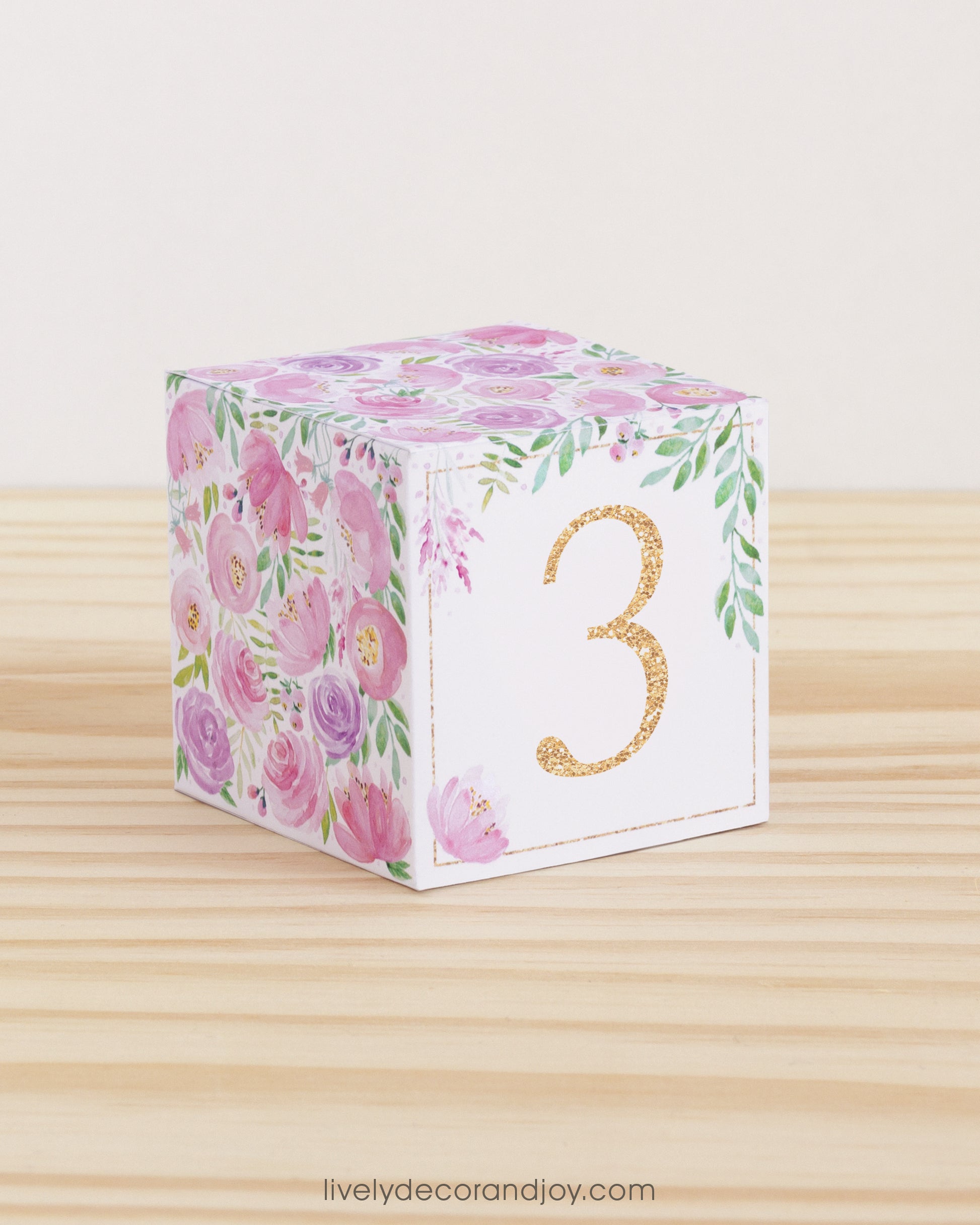 Paper cube with a big blush number 3 on the front and blush and lavender floral designs all around. It is on a wooden table.