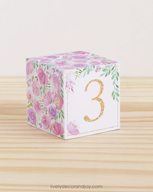Paper cube with a big blush number 3 on the front and blush and lavender floral designs all around. It is on a wooden table.