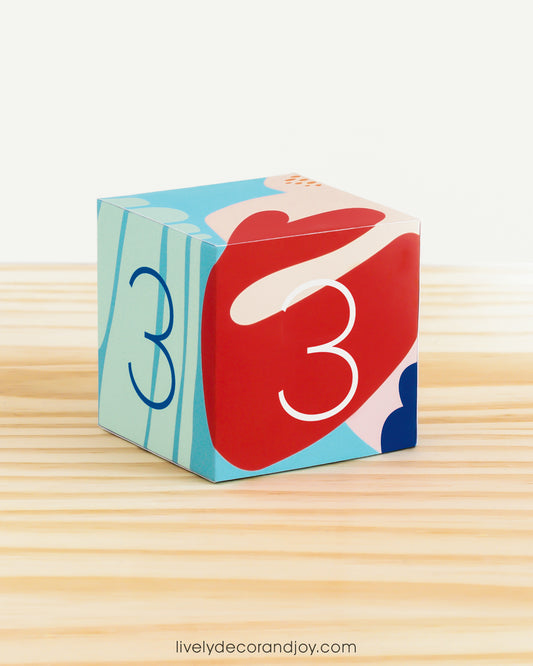 Paper building block in coral reef colors. It has the number 3 on the sides and is on a wood table or shelf.