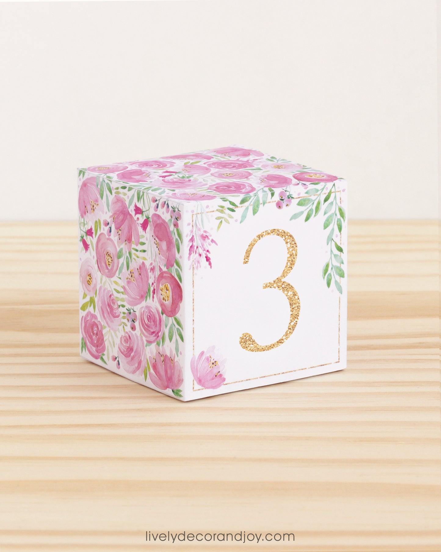 Paper cube with a big gold number 3 on the front and pink floral designs all around. It is on a wooden table.