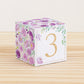 Paper cube with a big gold number 3 on the front and purple floral designs all around. It is on a wooden table.
