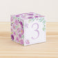 Paper cube with a big lavender number 3 on the front and purple floral designs all around. It is on a wooden table.