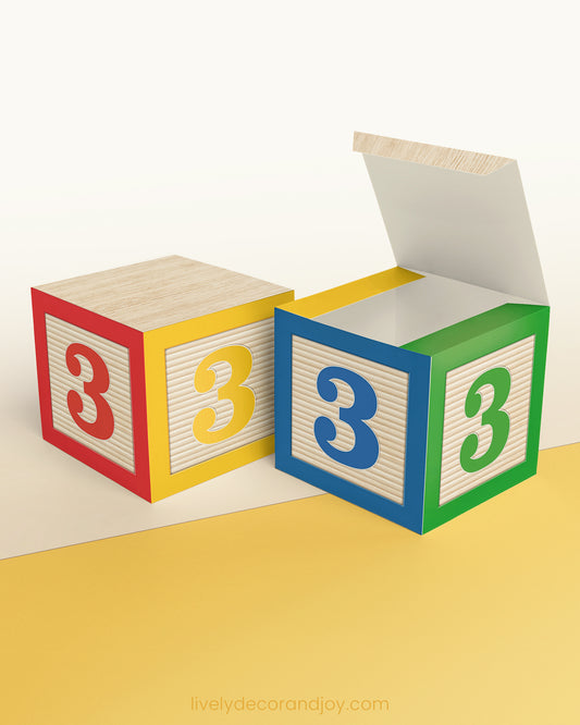 Paper building block in red, yellow, blue, and green. It has the number 3 on the sides and is on a wood table or shelf.