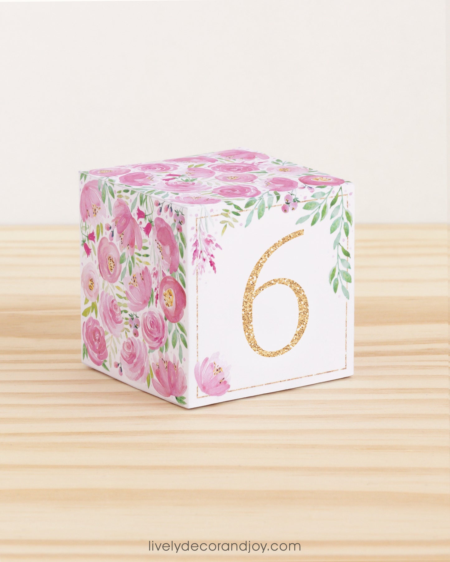 Paper building block with pink flowers, green leaves, and the digit 6 in gold. It is on a wooden table shelf or table.