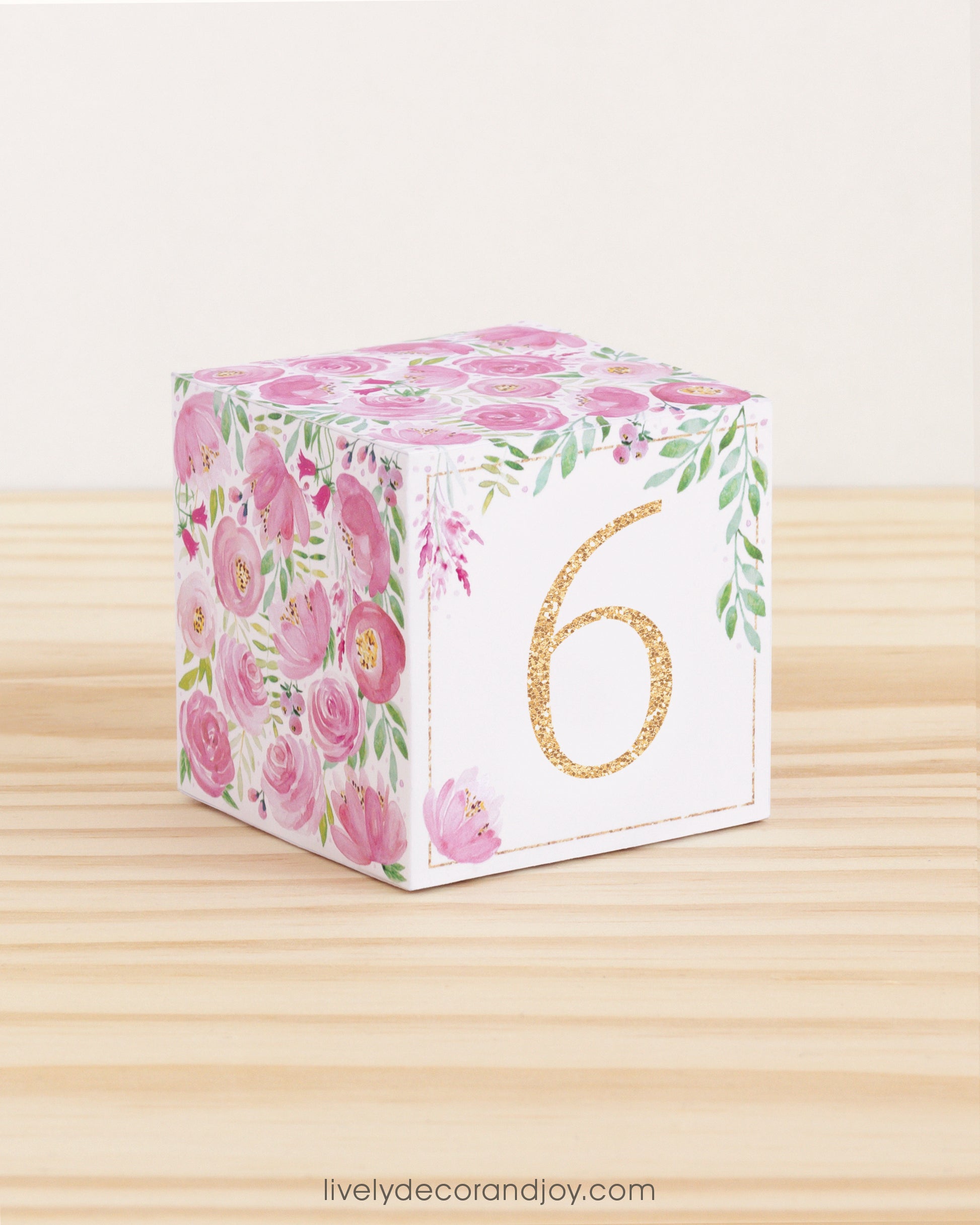 Paper building block with pink flowers, green leaves, and the digit 6 in gold. It is on a wooden table shelf or table.