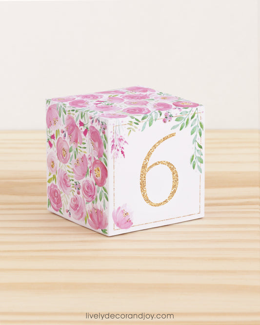 Paper building block with pink flowers, green leaves, and the digit 6 in gold. It is on a wooden table shelf or table.