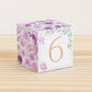 Paper building block with purple flowers, green leaves, and the digit 6 in gold. It is on a wooden table shelf or table.