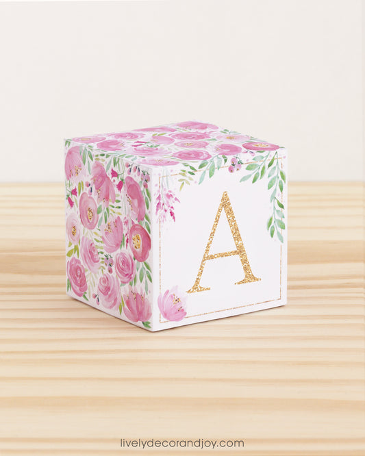 Large DIY block with the letter A in gold and floral designs in pink and green.The paper cube is on a wooden table.