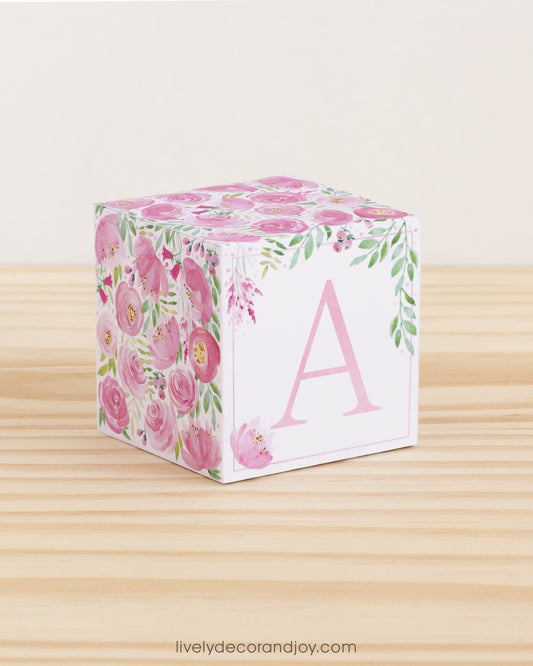 Large DIY block with the letter A in pink and floral designs in pink and green.The paper cube is on a wooden table.