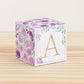Large DIY block with the letter A in gold and floral designs in purple and green.The paper cube is on a wooden table.