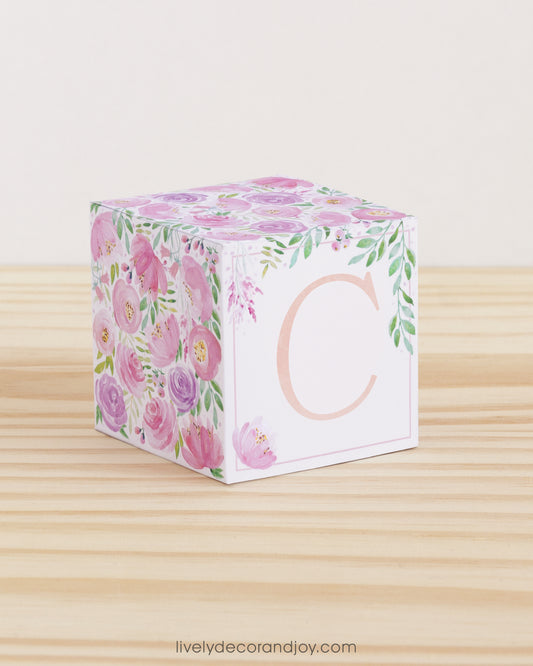 A paper cube with boho designs on a wooden surface. On the front, it has a capital C and a thin square frame in peach.