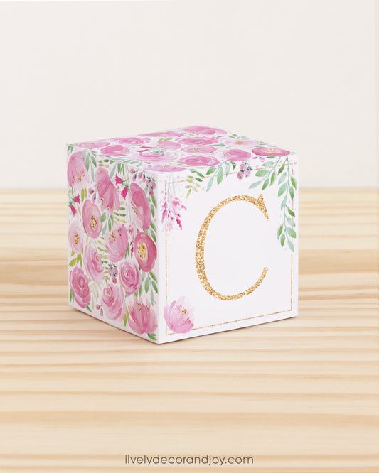 A paper cube with boho designs on a wooden surface. On the front, it has a capital C and a thin square frame in gold.