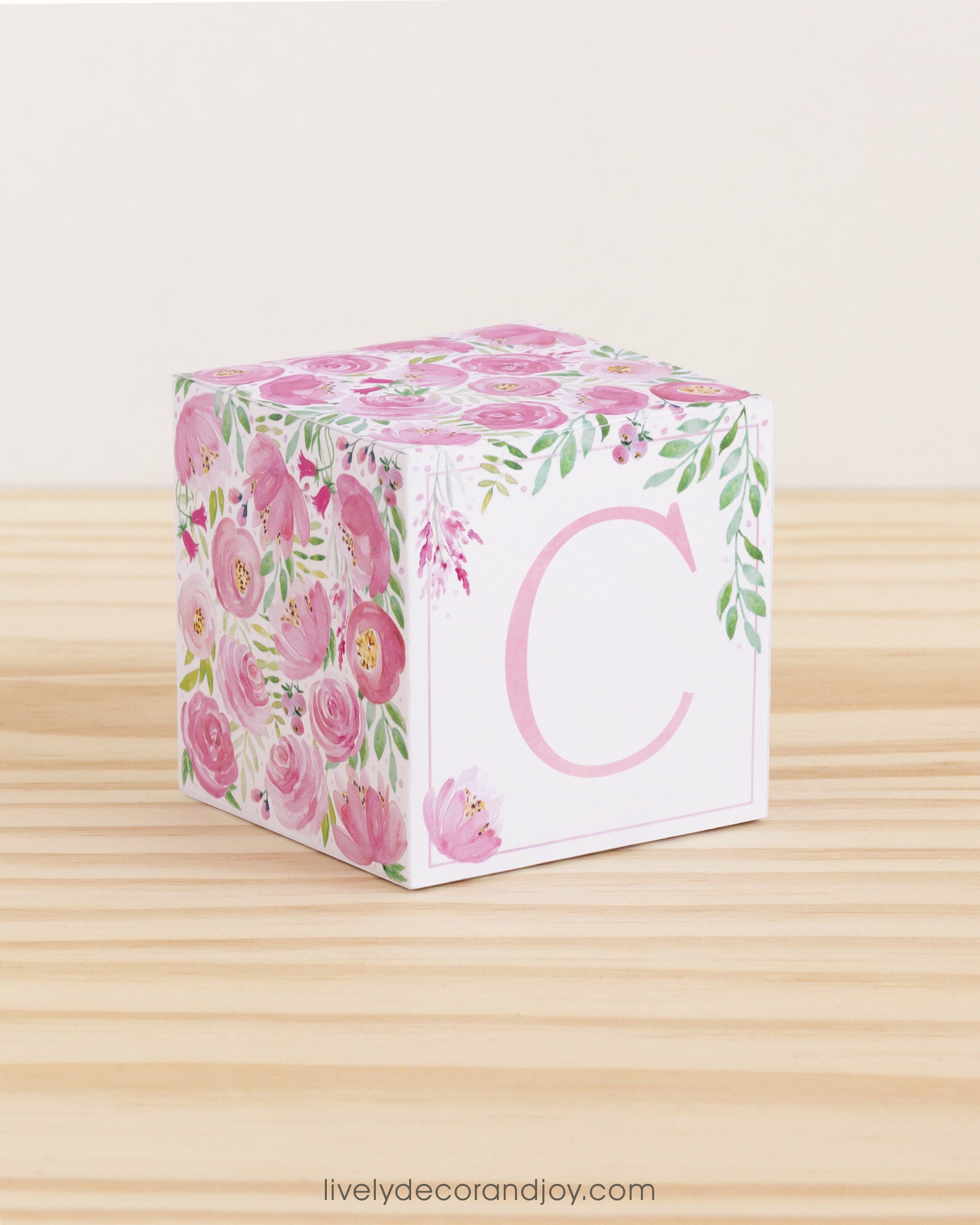A paper cube with boho designs on a wooden surface. On the front, it has a capital C and a thin square frame in pink.