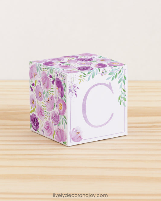 A paper cube with boho designs on a wooden surface. On the front, it has a capital C and a thin square frame in lavender.