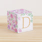 On a wooden table is a paper building block with blush and lavender floral designs and the letter D in peach on the front.