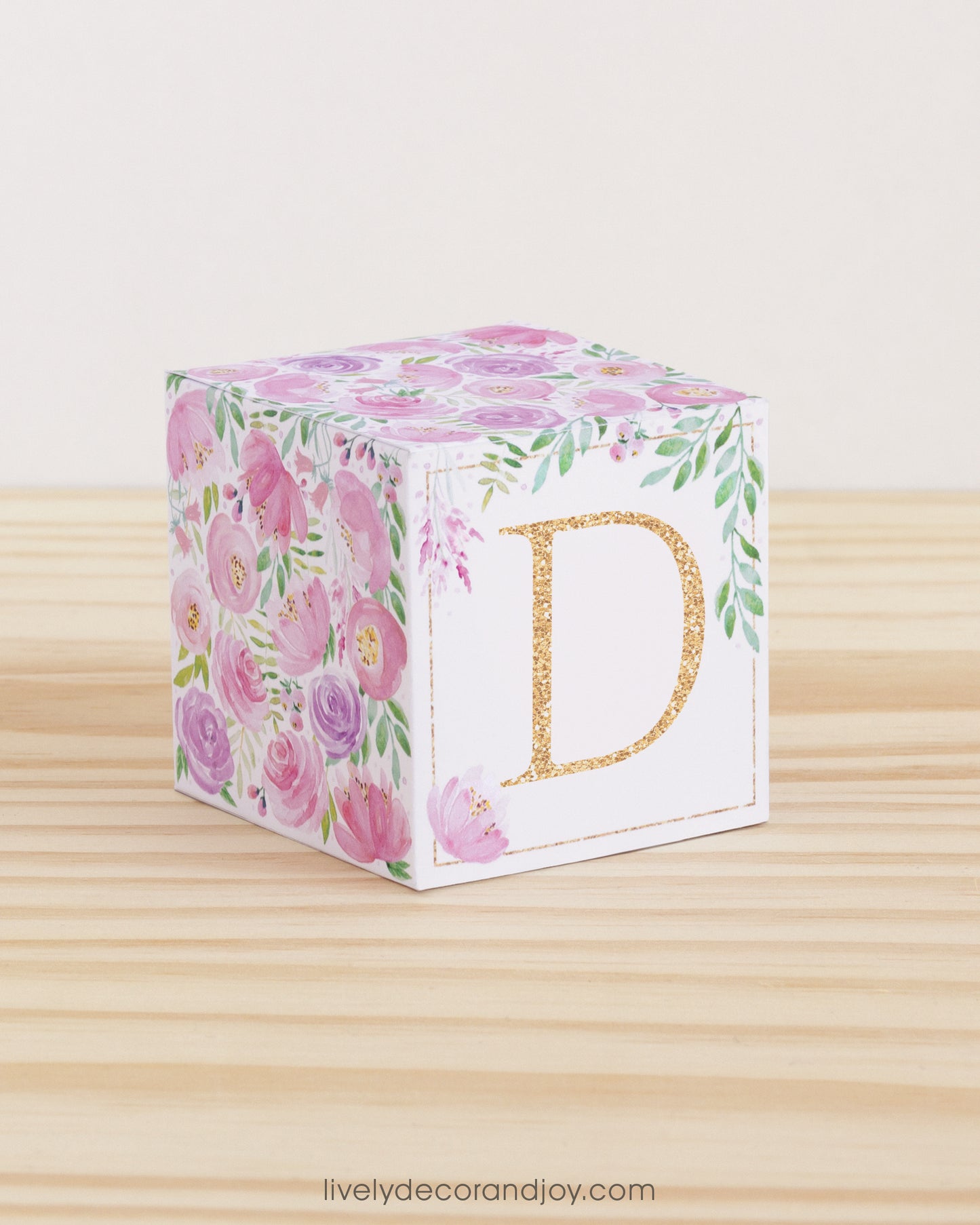 On a wooden table is a paper building block with blush and lavender floral designs and the letter D in peach on the front.