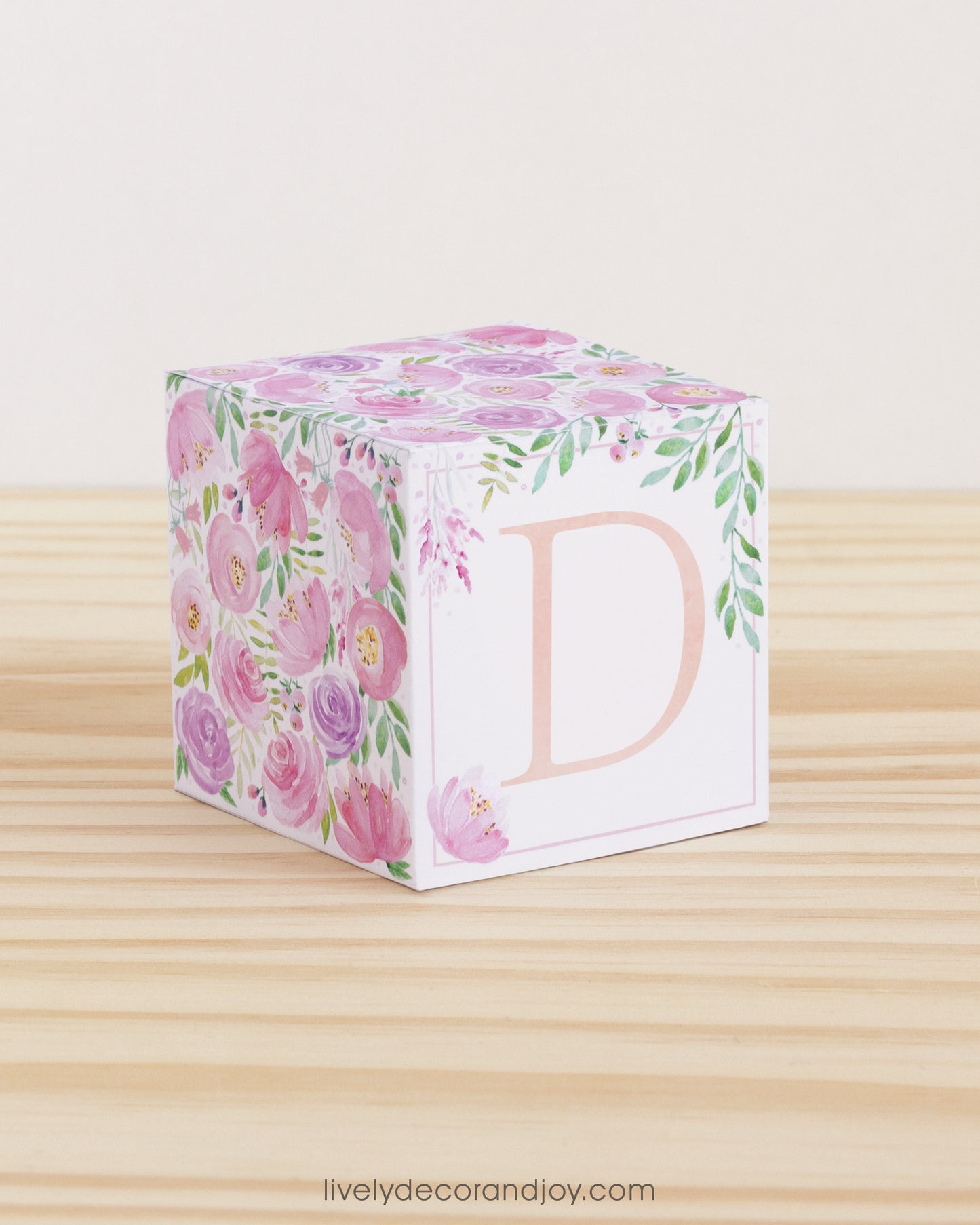 On a wooden table is a paper building block with blush and lavender floral designs and the letter D in peach on the front.