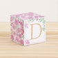 On a wooden table is a paper building block with pink and green floral designs and the letter D in gold on the front.