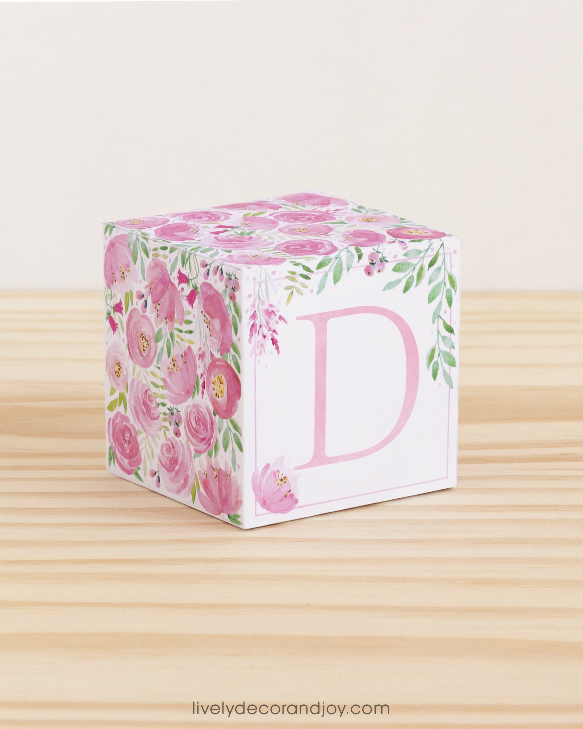 On a wooden table is a paper building block with pink and green floral designs and the letter D in pink on the front.
