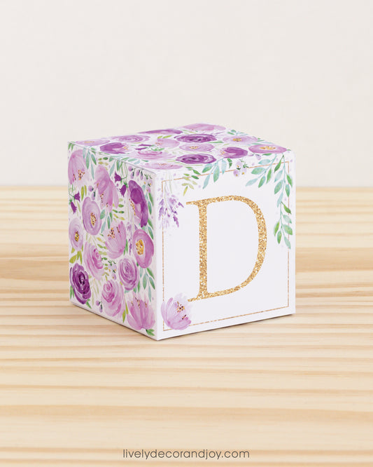 On a wooden table is a paper building block with purple and green floral designs and the letter D in gold on the front.