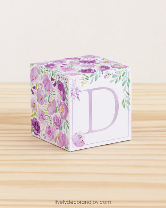 On a wooden table is a paper building block with purple and green floral designs and the letter D in lavender on the front.