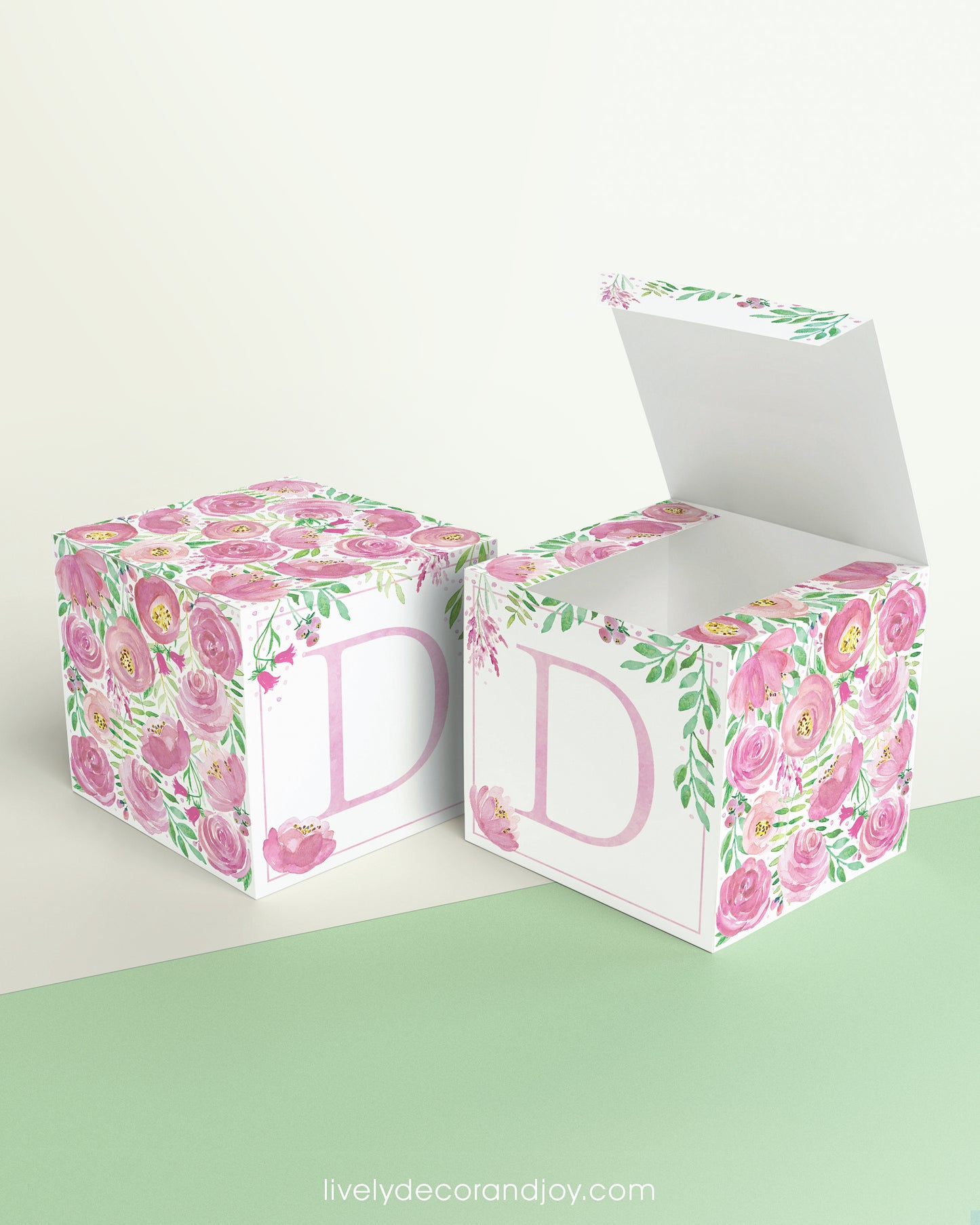 Two paper cubes with floral designs and the letter D in pink. One has the top open, showing it can be a cube or a box.