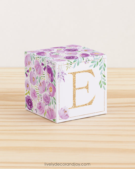 A baby block with purple and green floral designs and a gold letter E is on a wooden surface. It is a paper building block.