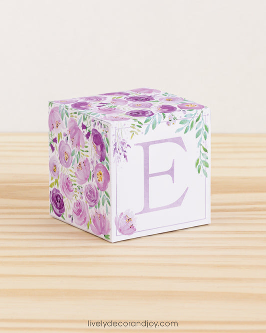 Baby block with purple and green floral designs and lavender letter E is on a wooden surface. It is a paper building block.