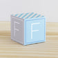 ABC block in blue and light grey with the letter F on the sides. It is on a wooden surface.
