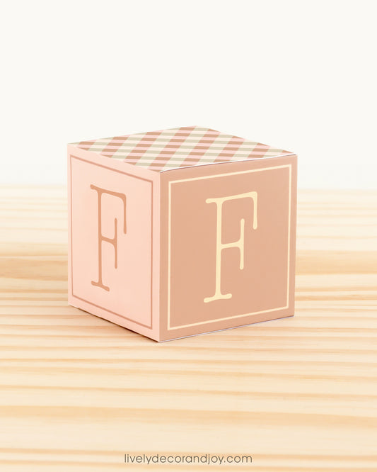 ABC block in olive green, pale pink and brown with the letter F on the sides. It is on a wooden surface.