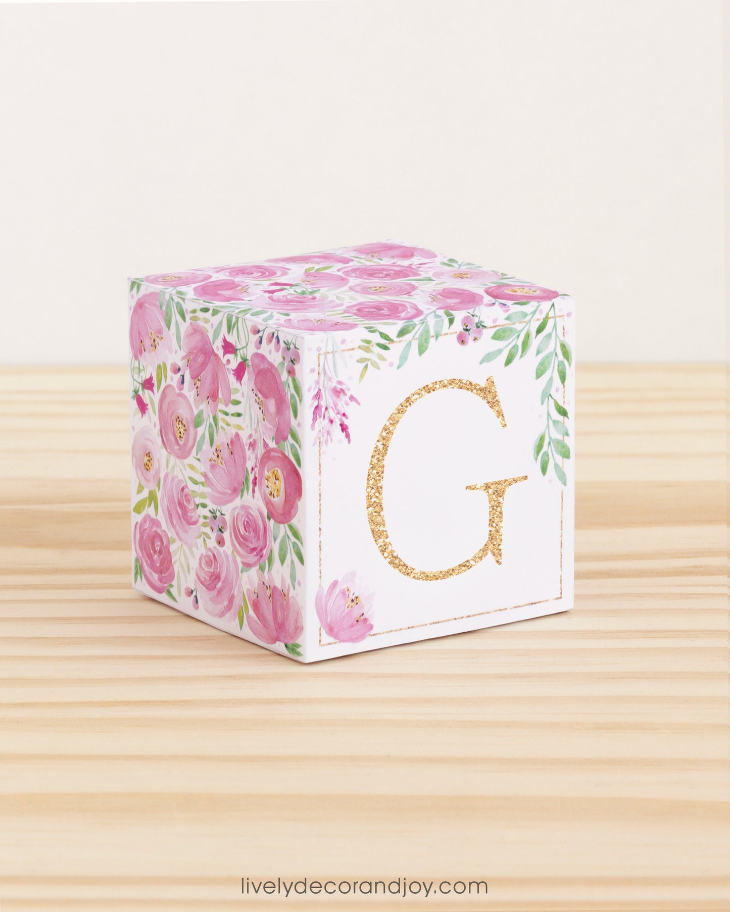 Letter block with printed pink and green floral designs and a gold capital G on the front. It is made of paper.
