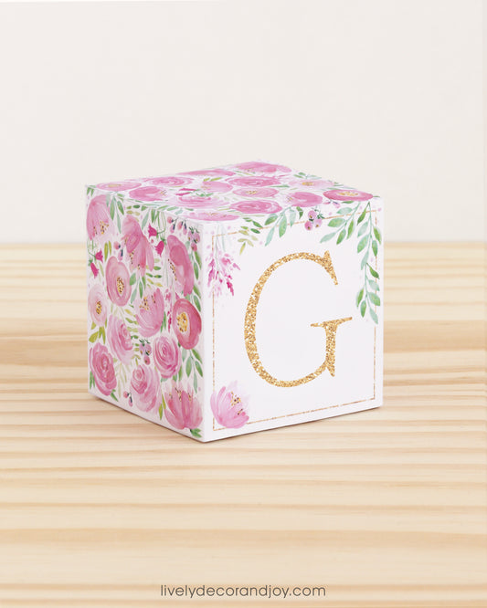 Letter block with printed pink and green floral designs and a gold capital G on the front. It is made of paper.