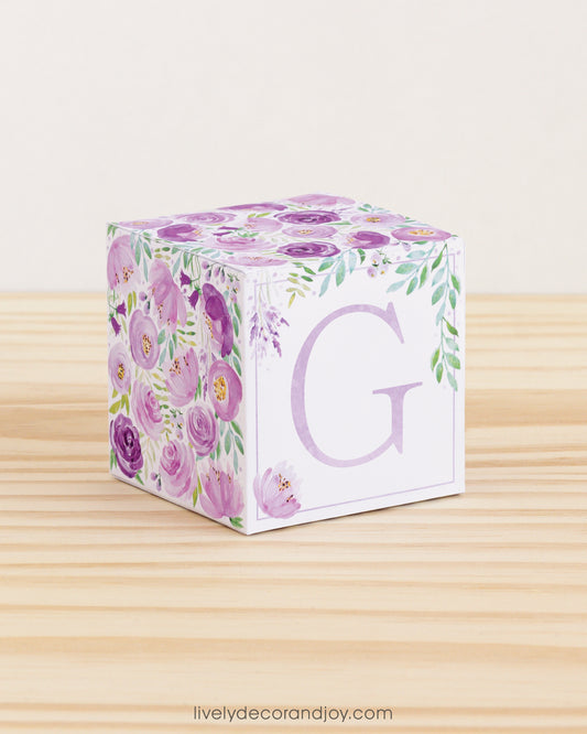 Letter block with printed purple and green floral designs and a lavender capital G on the front. It is made of paper.