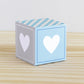 Paper alphabet cube with a heart on the sides. Designed in tints of blue and light grey. It is on a wooden table.