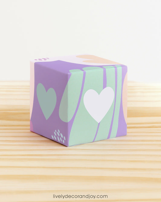 Paper alphabet cube with a heart on the sides. Designed in pastel colors. It is on a wooden table.