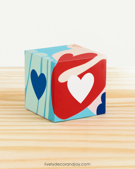 Paper alphabet cube with a heart on the sides. Designed in coral reef colors. It is on a wooden table.