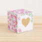 Paper cube with a big gold heart on the front and pink floral designs all around. It is on a wooden table.
