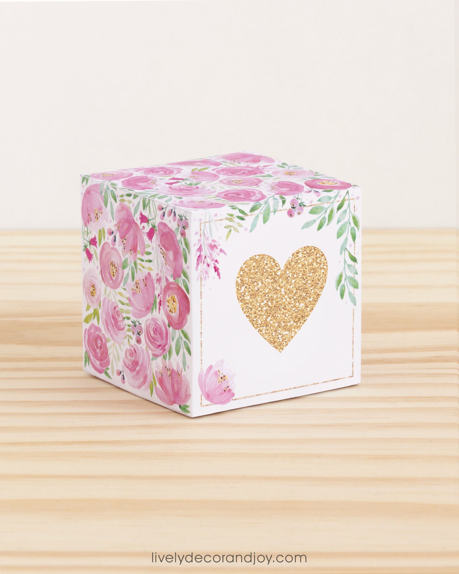 Paper cube with a big gold heart on the front and pink floral designs all around. It is on a wooden table.