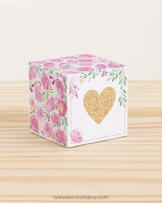 Paper cube with a big gold heart on the front and pink floral designs all around. It is on a wooden table.