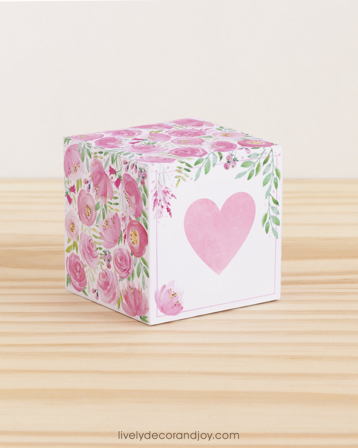 Paper cube with a big pink heart on the front and pink floral designs all around. It is on a wooden table.