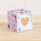 Paper cube with a big gold heart on the front and purple floral designs all around. It is on a wooden table.