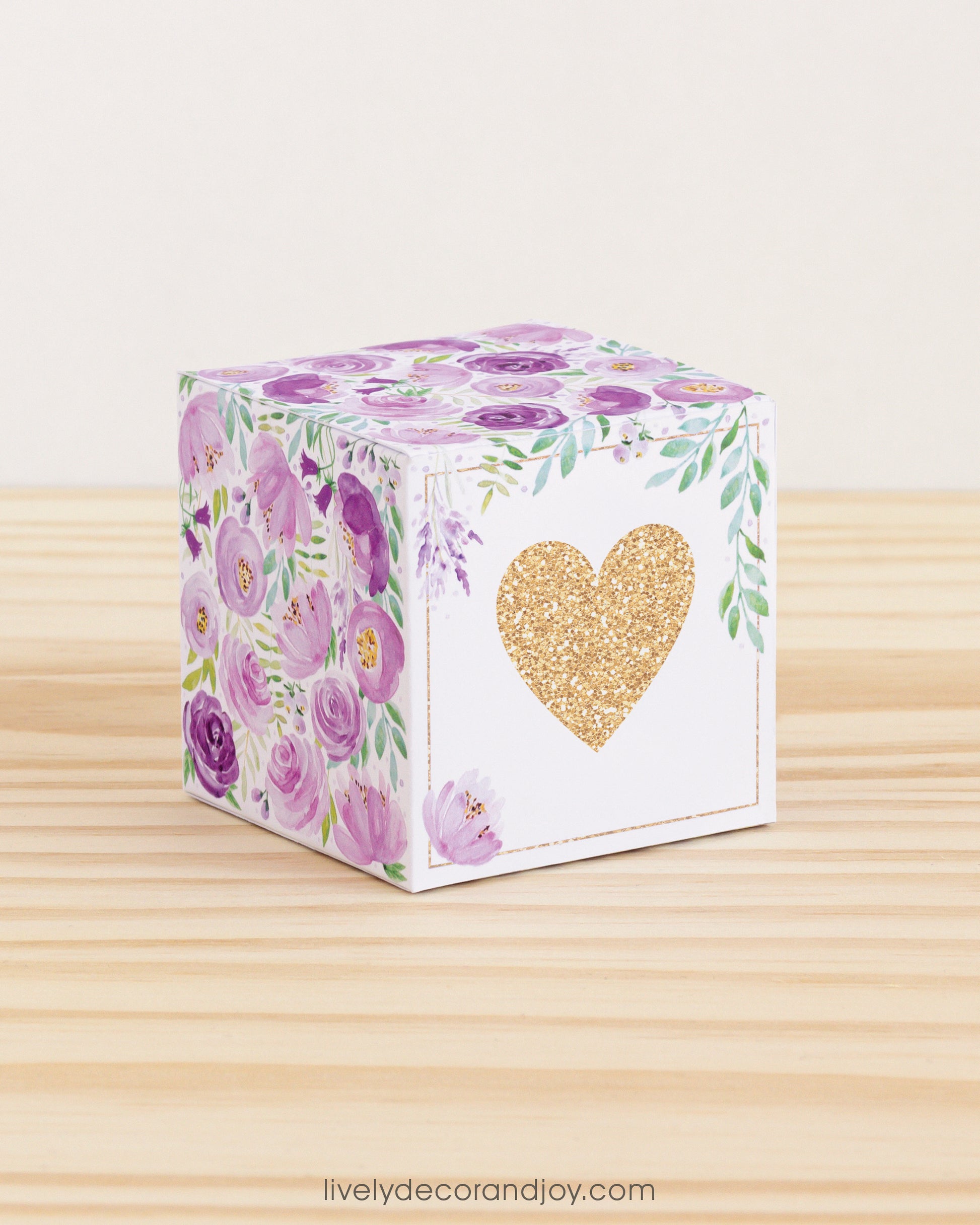 Paper cube with a big gold heart on the front and purple floral designs all around. It is on a wooden table.