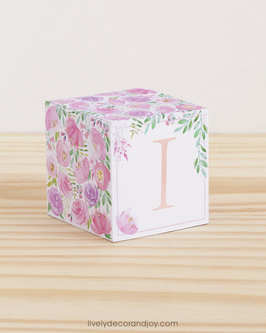 Building block made of printed paper with floral designs and the letter I on the front. It is on a wooden table.