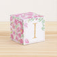 Building block made of printed paper with floral designs and the letter I on the front. It is on a wooden table.