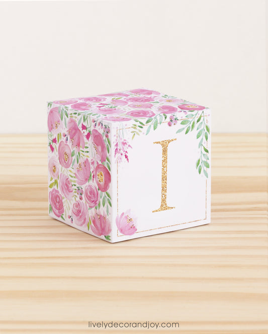 Building block made of printed paper with floral designs and the letter I on the front. It is on a wooden table.