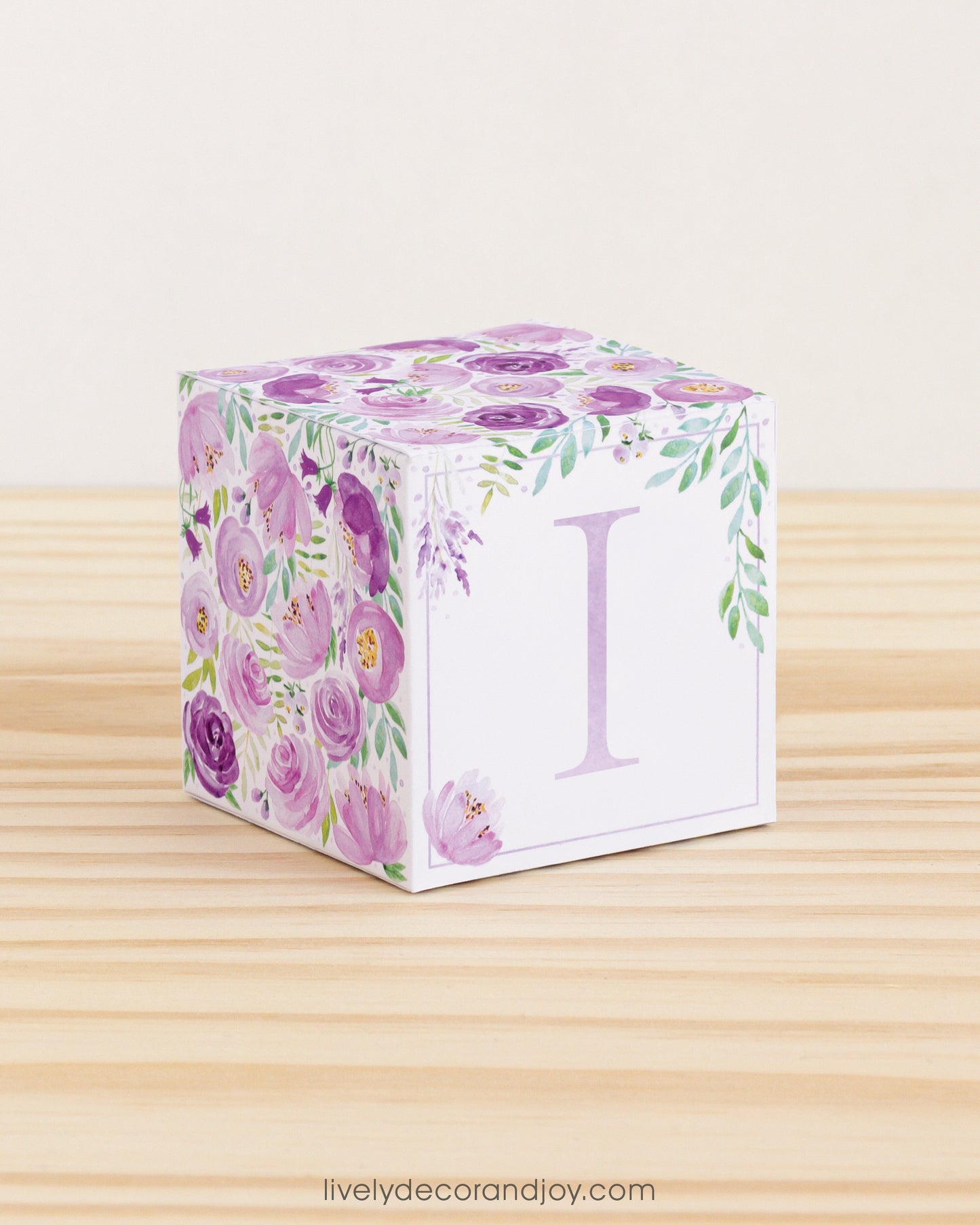 Building block made of printed paper with floral designs and the letter I on the front. It is on a wooden table.