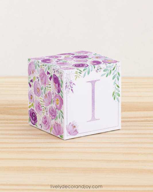 Building block made of printed paper with floral designs and the letter I on the front. It is on a wooden table.