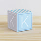 Big DIY block with the letter K in tints of blue and light grey.The paper cube is on a wooden table.
