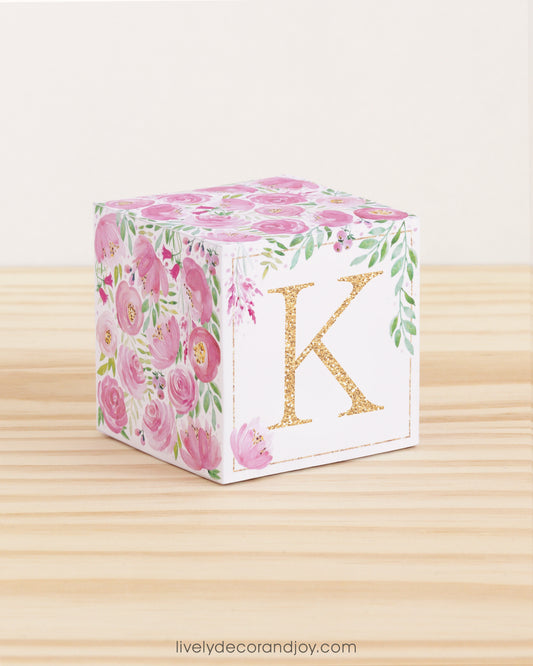 Big DIY block with the letter K in gold and floral designs in pink and green.The paper cube is on a wooden table.