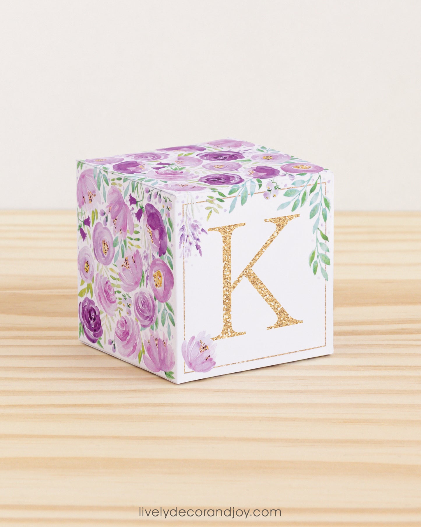 Big DIY block with the letter K in gold and floral designs in purple and green.The paper cube is on a wooden table.